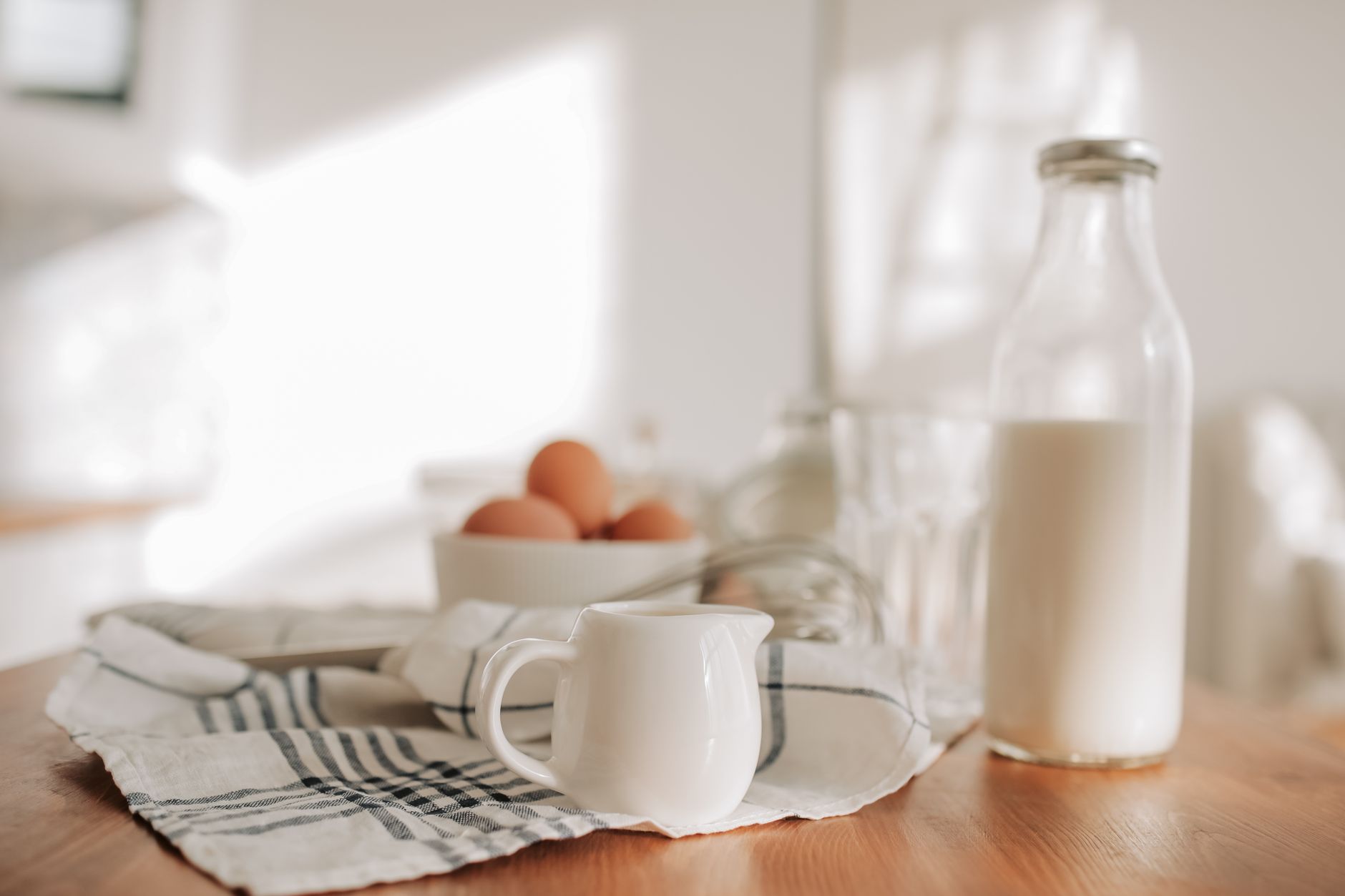 A clear glass bottle of milk on a wooden table, beside a small white jug, a bowl of brown eggs, and a blue-striped cloth.