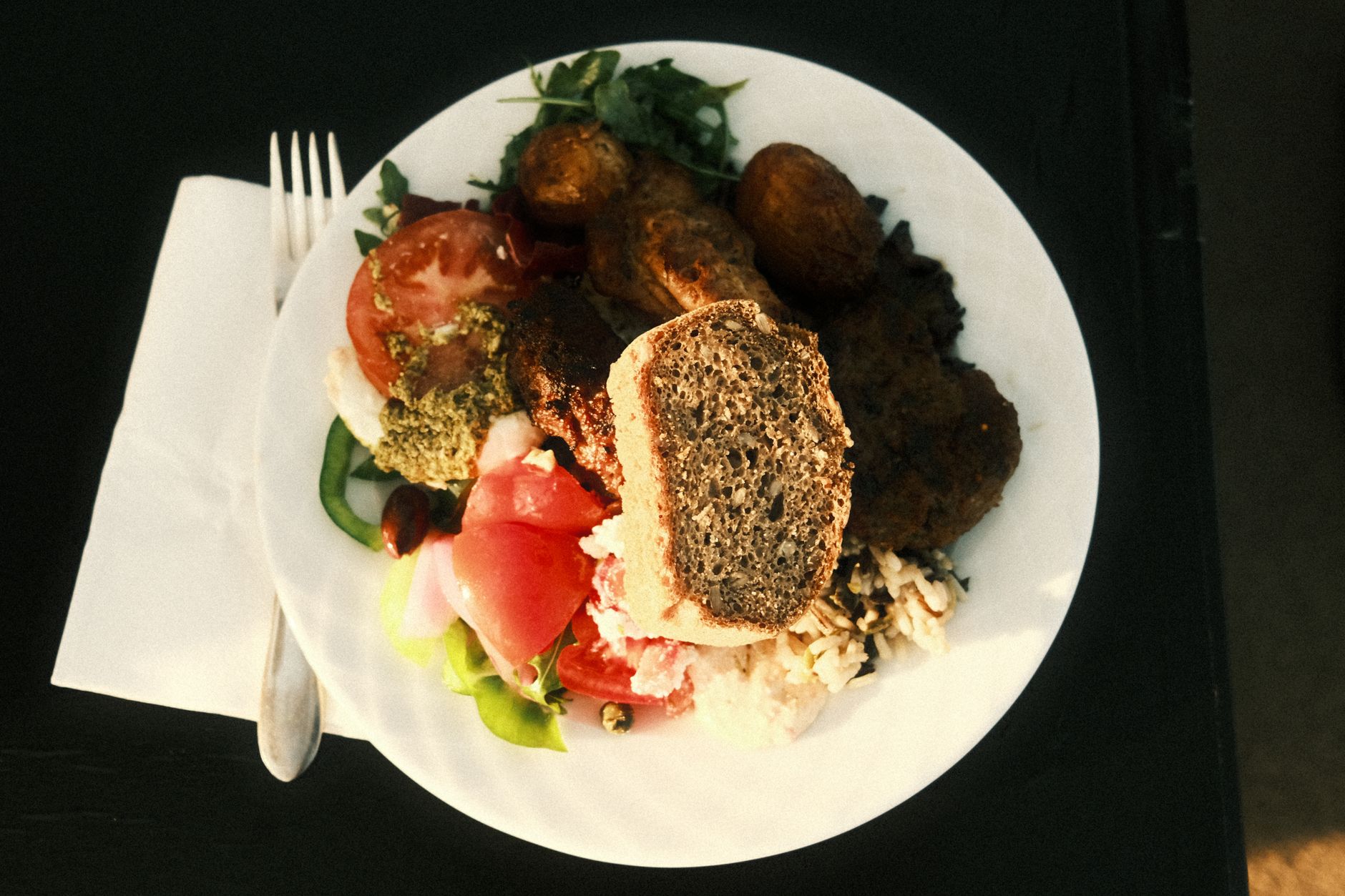 A white plate from above holds tomato and cucumber salad, rice, a slice of seeded bread, and browned pieces of food, with a fork on a white napkin beside it.