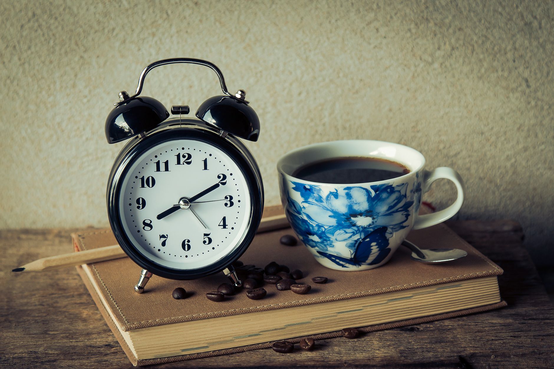 A black twin-bell alarm clock stands on a closed tan book beside a blue and white cup of coffee, a pencil, and a few coffee beans on a wooden table.