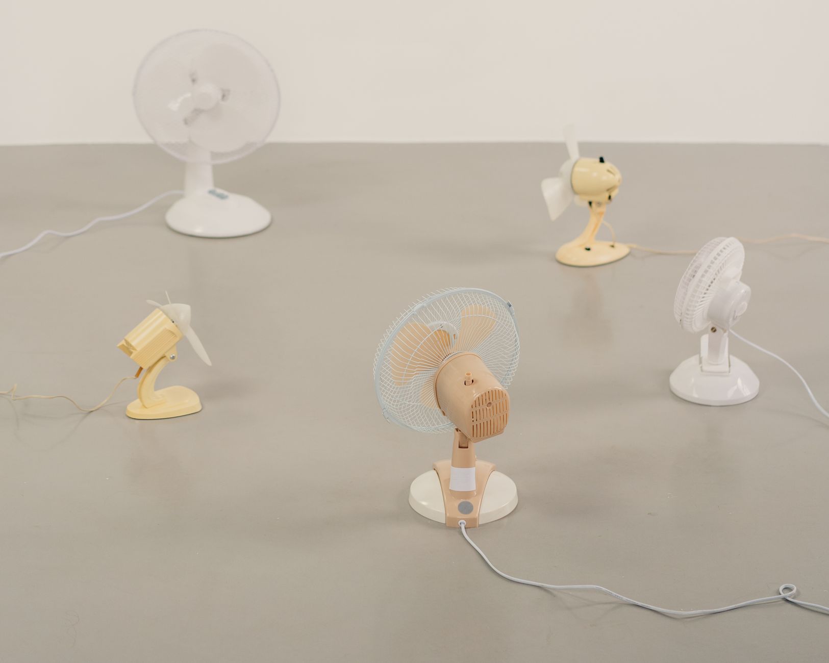 Five small desk fans in white, cream and pale wood, with their cords, arranged on a plain grey floor.