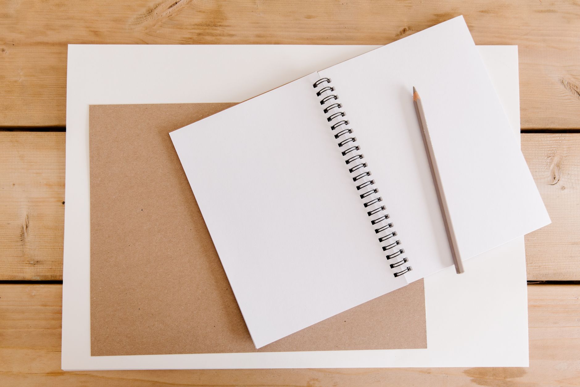A blank spiral notebook and a grey pencil lying on white paper, over a brown card, on a pale wooden table.