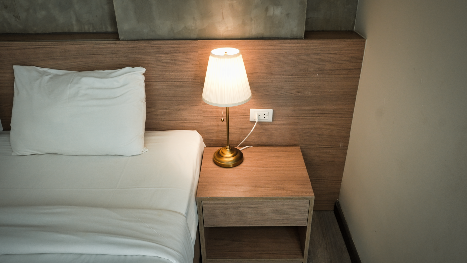 A white pillow and sheet on a made bed beside a wooden nightstand, with a brass lamp switched on against a wood-panelled wall.
