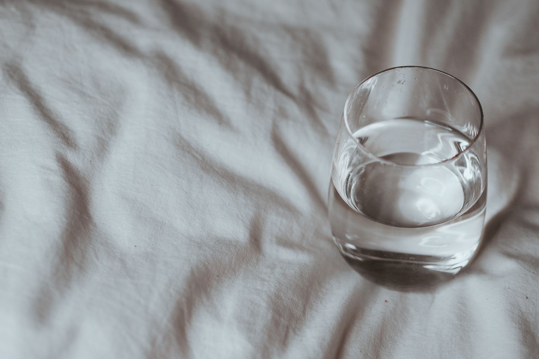 A short clear glass of water on rumpled white bed linen, with soft light and no other objects.