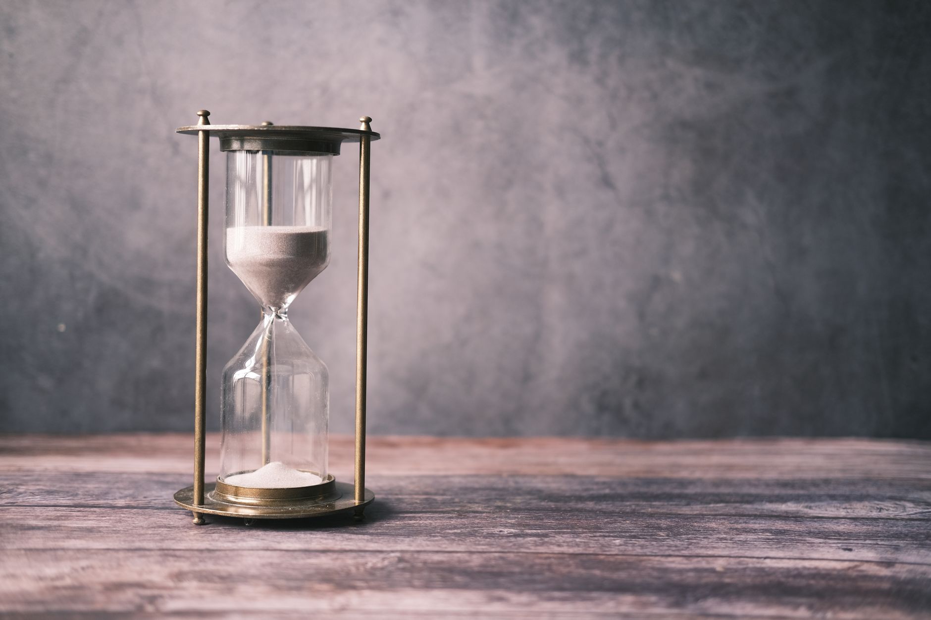 A brass-framed hourglass with white sand in both glass bulbs stands on a weathered wooden table against a plain grey wall.