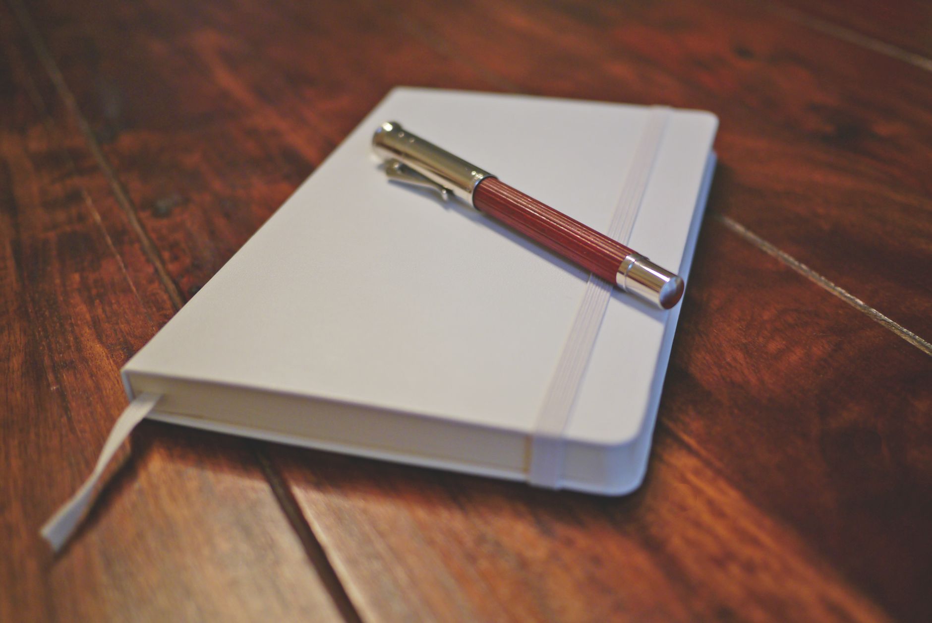 A closed white notebook with a pale elastic band lies on a wooden table, with a red and silver pen resting across the cover.