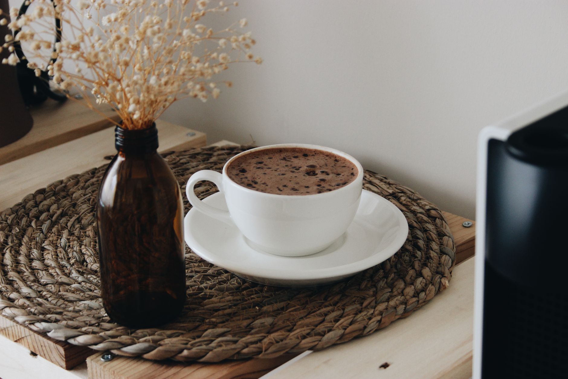 A brown glass bottle holding dried flowers stands beside a white cup and saucer of coffee on a round woven mat.