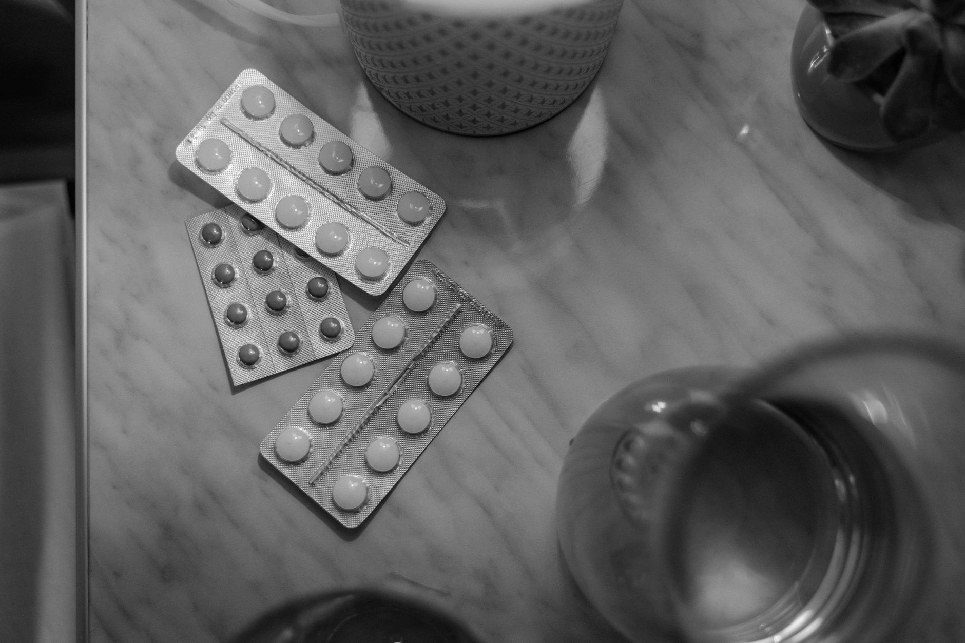 Three silver blister packs of round tablets lie on a pale marble table beside a dotted cup and the rim of a glass of water, photographed in black and white.