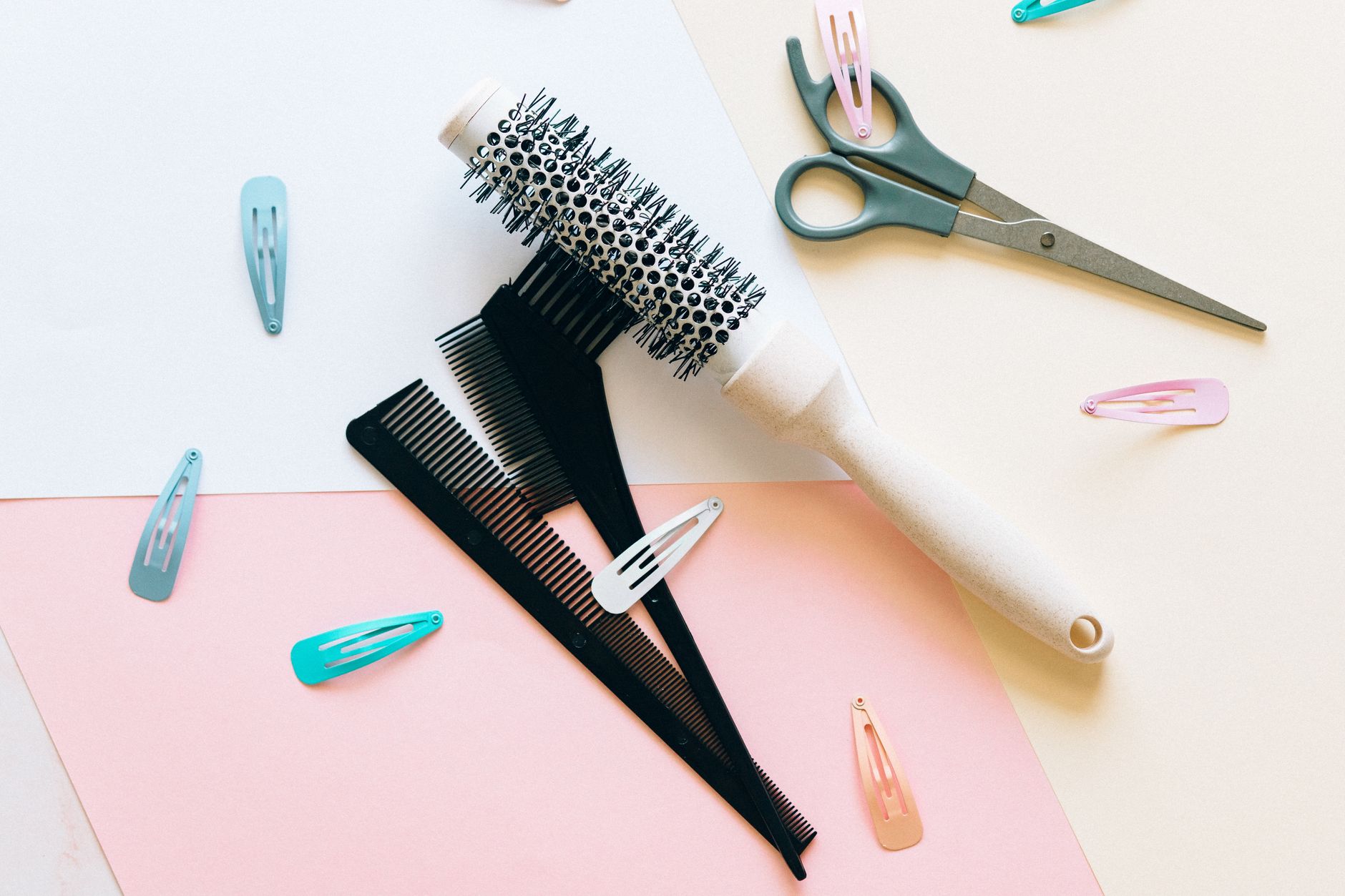 A round hairbrush, a black comb and small scissors laid on pink, white and cream paper, with a few pastel hair clips around them.