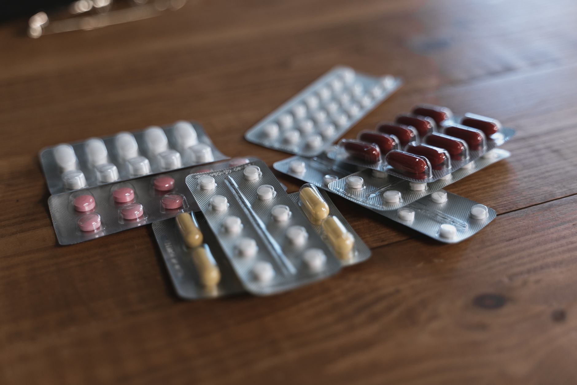 Several silver blister packs of white, pink, yellow and dark red tablets and capsules overlap on a wooden table.