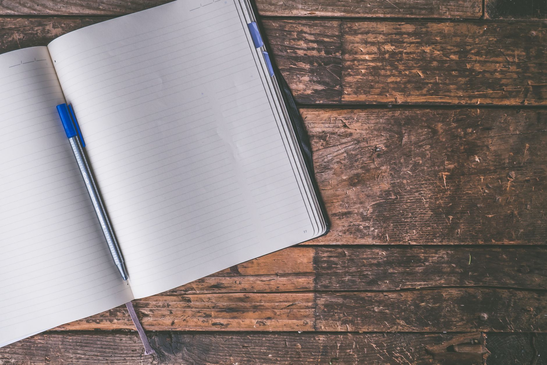 An open lined notebook on a weathered wooden table, with a blue and silver pen lying on the left page.