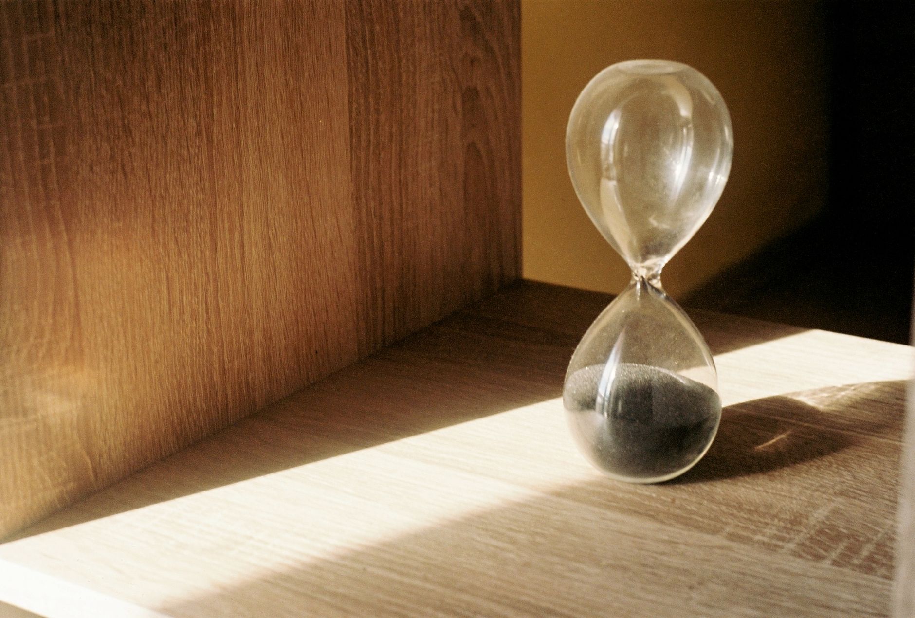 A clear glass hourglass with black sand in the lower bulb, standing on a pale wooden shelf in a stripe of sunlight.