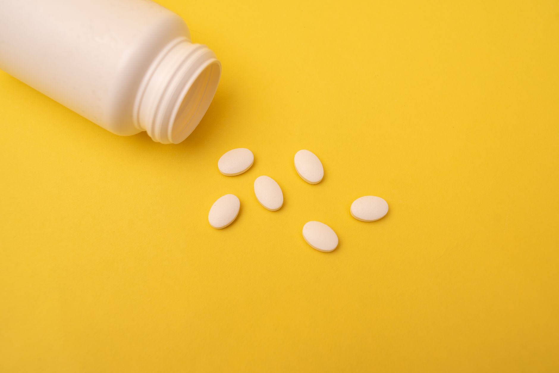 A white plastic bottle lying on its side on a yellow surface, with six white oval tablets spilled beside the open neck.