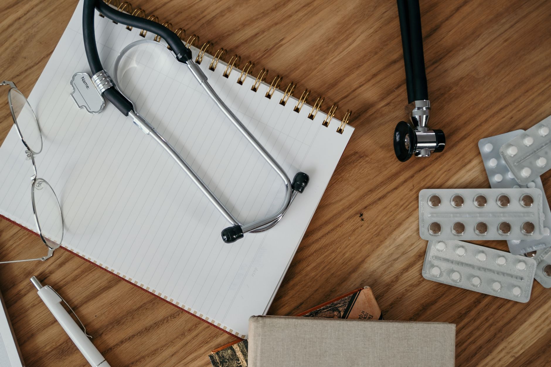 A stethoscope and wire-rimmed glasses lie on a blank spiral notebook, next to blister packs of tablets and a pen, on a wooden desk.
