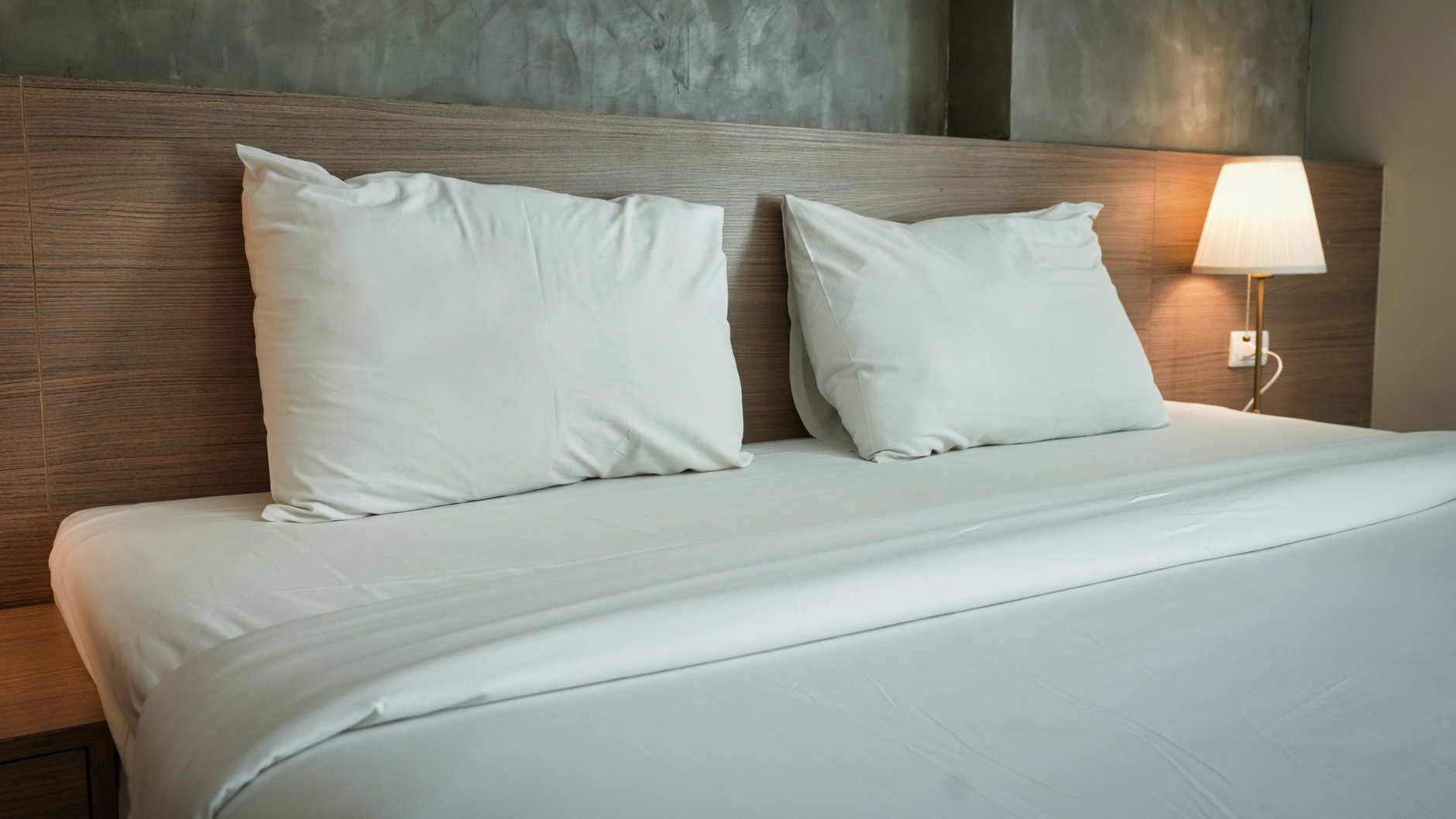 A made bed with two white pillows and a white duvet against a wooden headboard, and a lit wall lamp on the right.