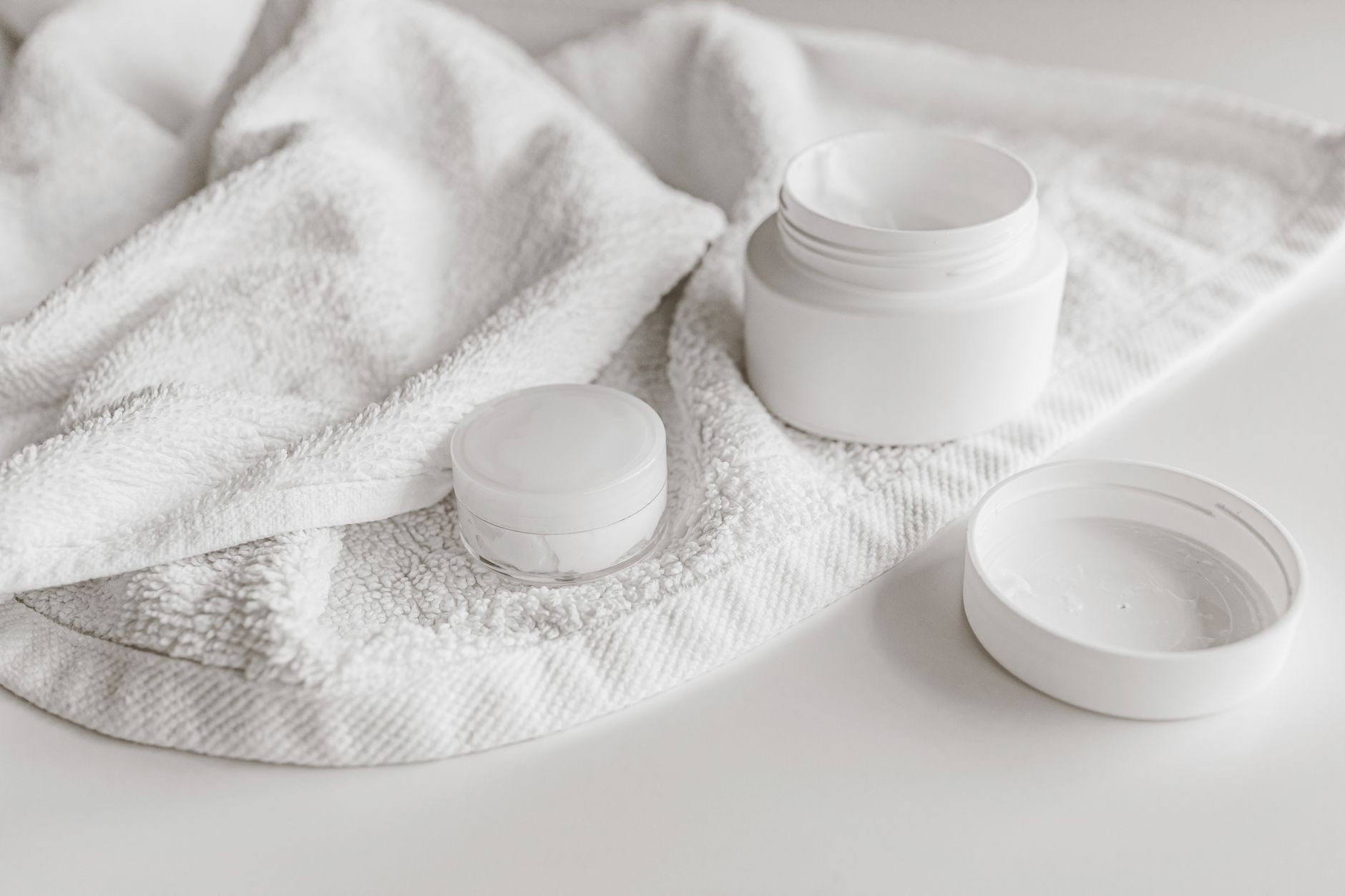 A white towel with an open jar of white cream, a small closed white jar, and a white lid on a pale surface.