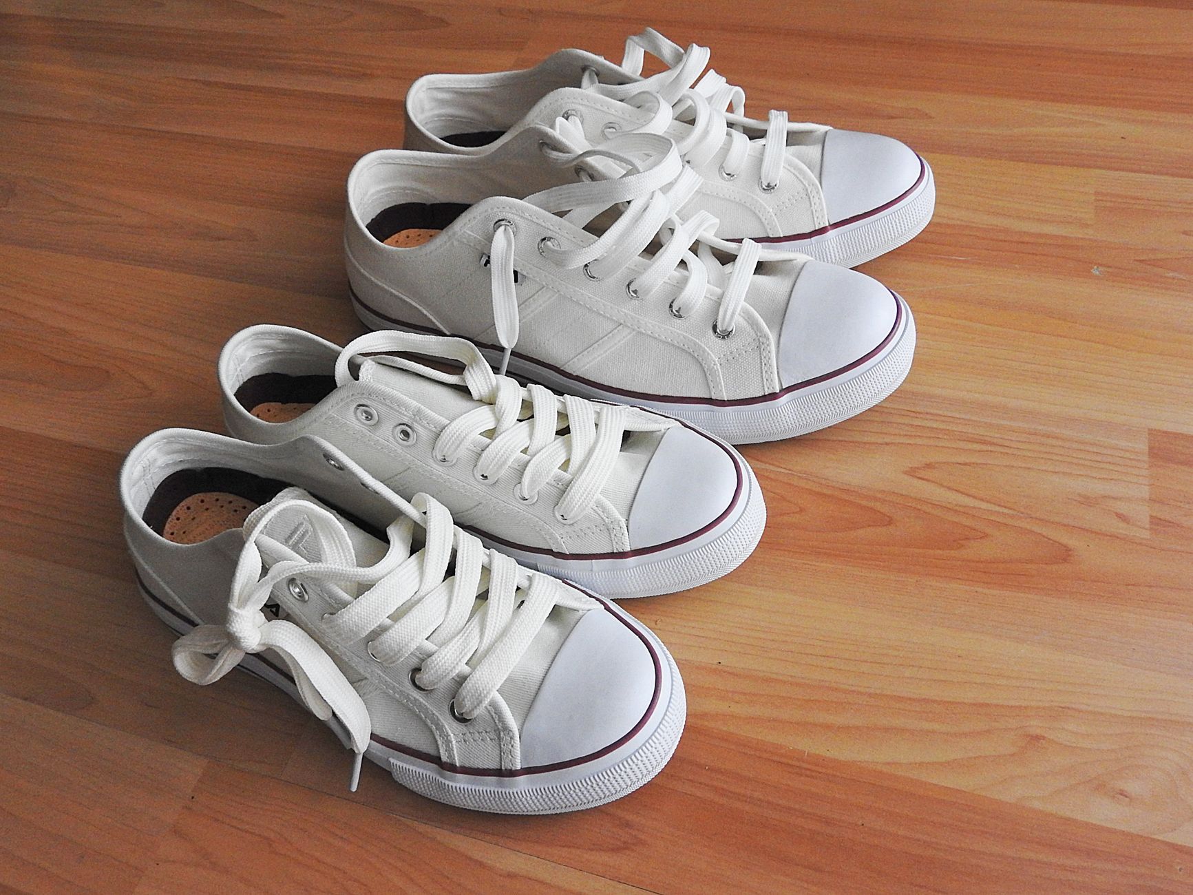 Two pairs of white canvas sneakers with white laces on a light wooden floor, one pair set slightly behind the other.