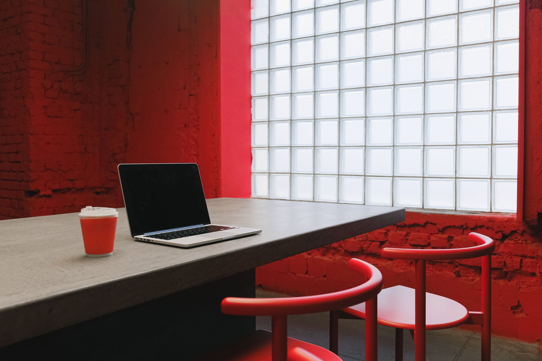 A grey table against a red brick wall, with an open silver laptop showing a dark screen, an orange paper cup, and two red chairs, beside a glass-block window.