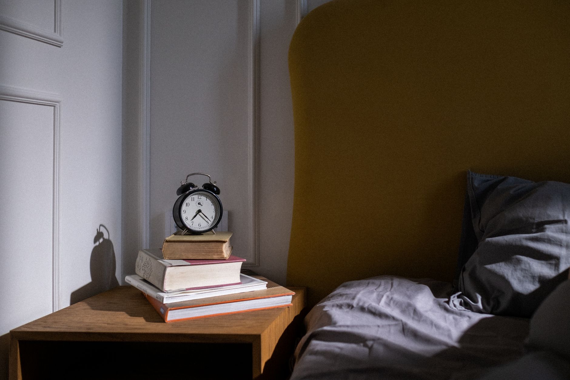 A black alarm clock on a stack of books on a wooden bedside table, next to a mustard headboard and grey bedding.