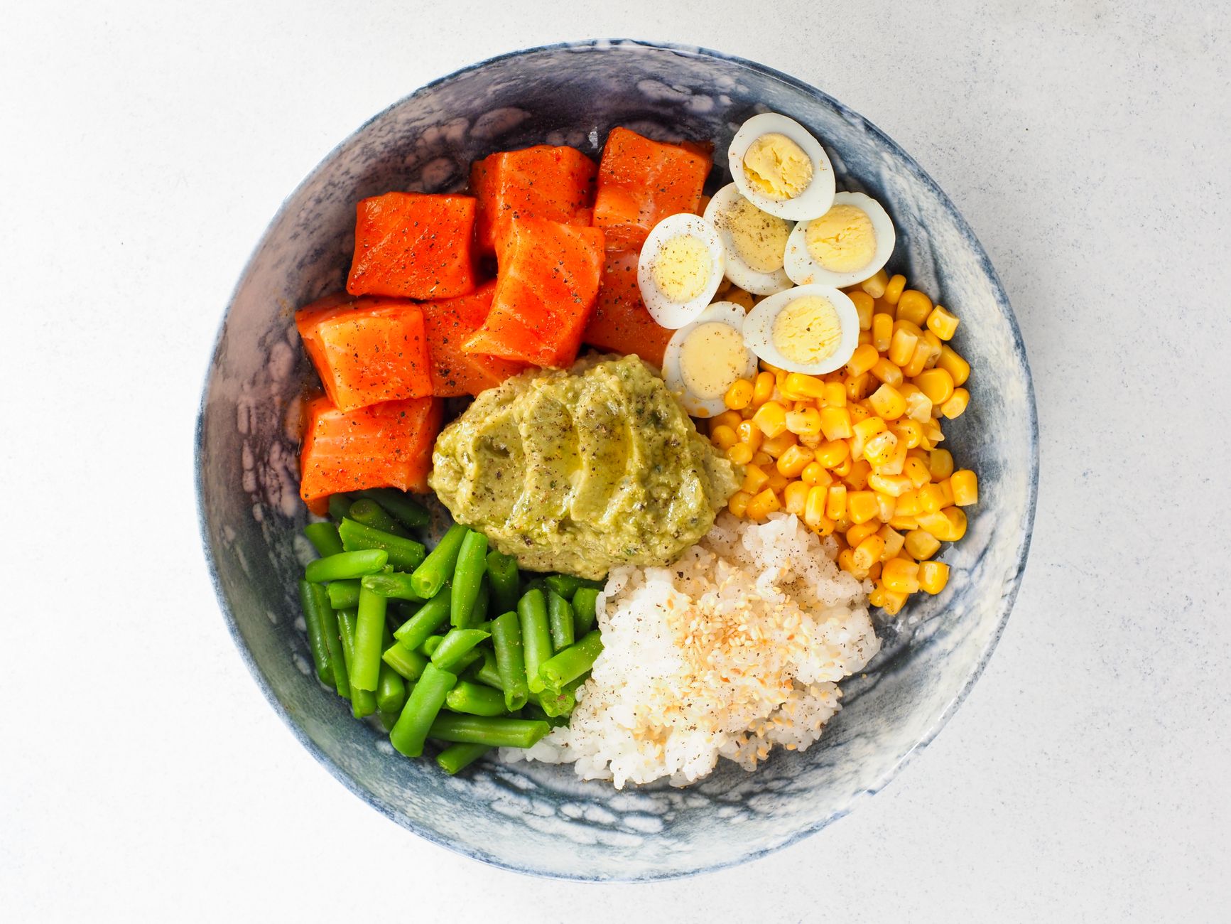 A speckled bowl seen from above holds cubed salmon, sliced boiled eggs, green beans, sweetcorn, rice and a green mash.