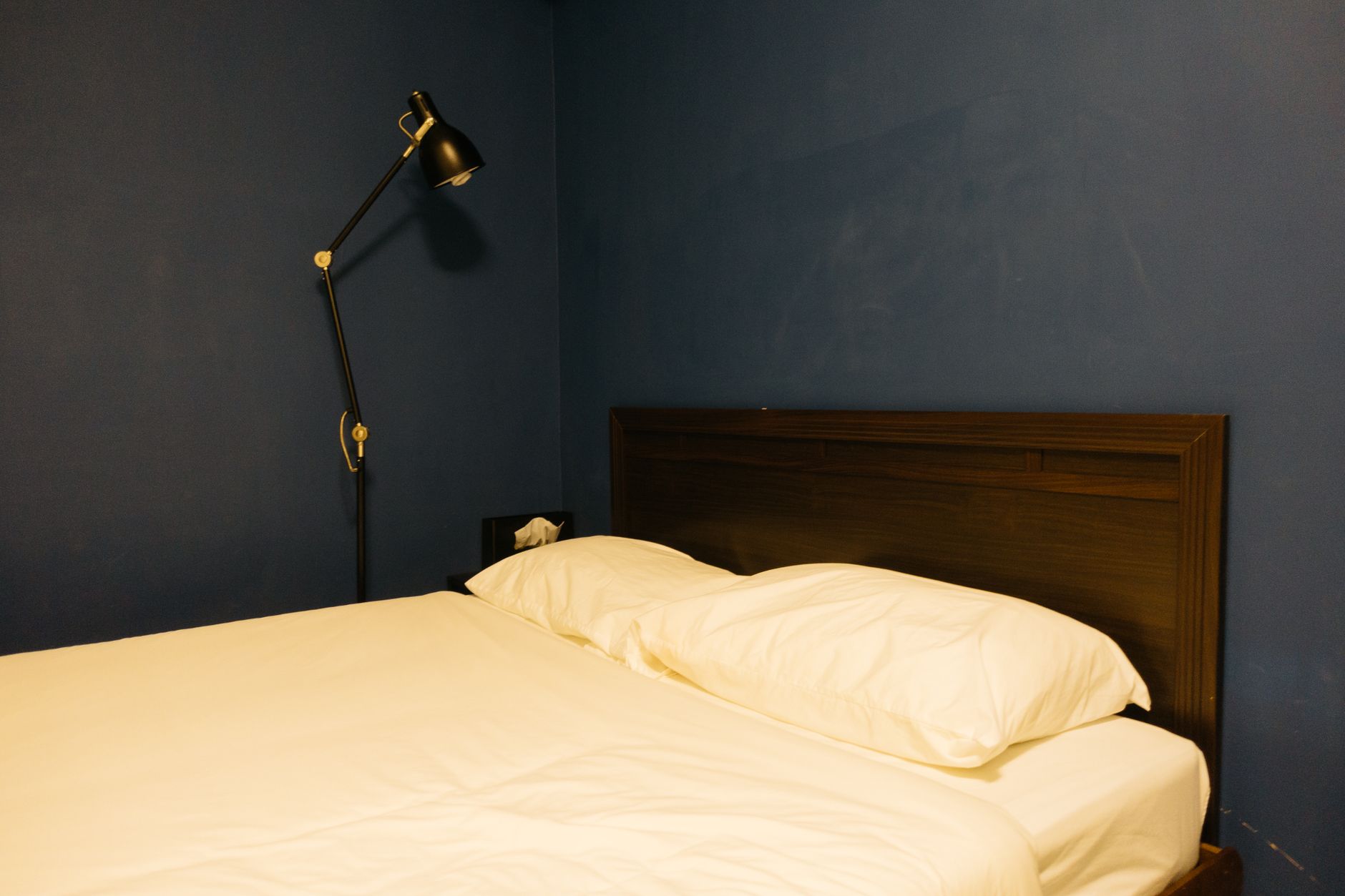 An empty bed with cream sheets and two white pillows against a dark wood headboard, beside a black adjustable lamp on a navy wall.