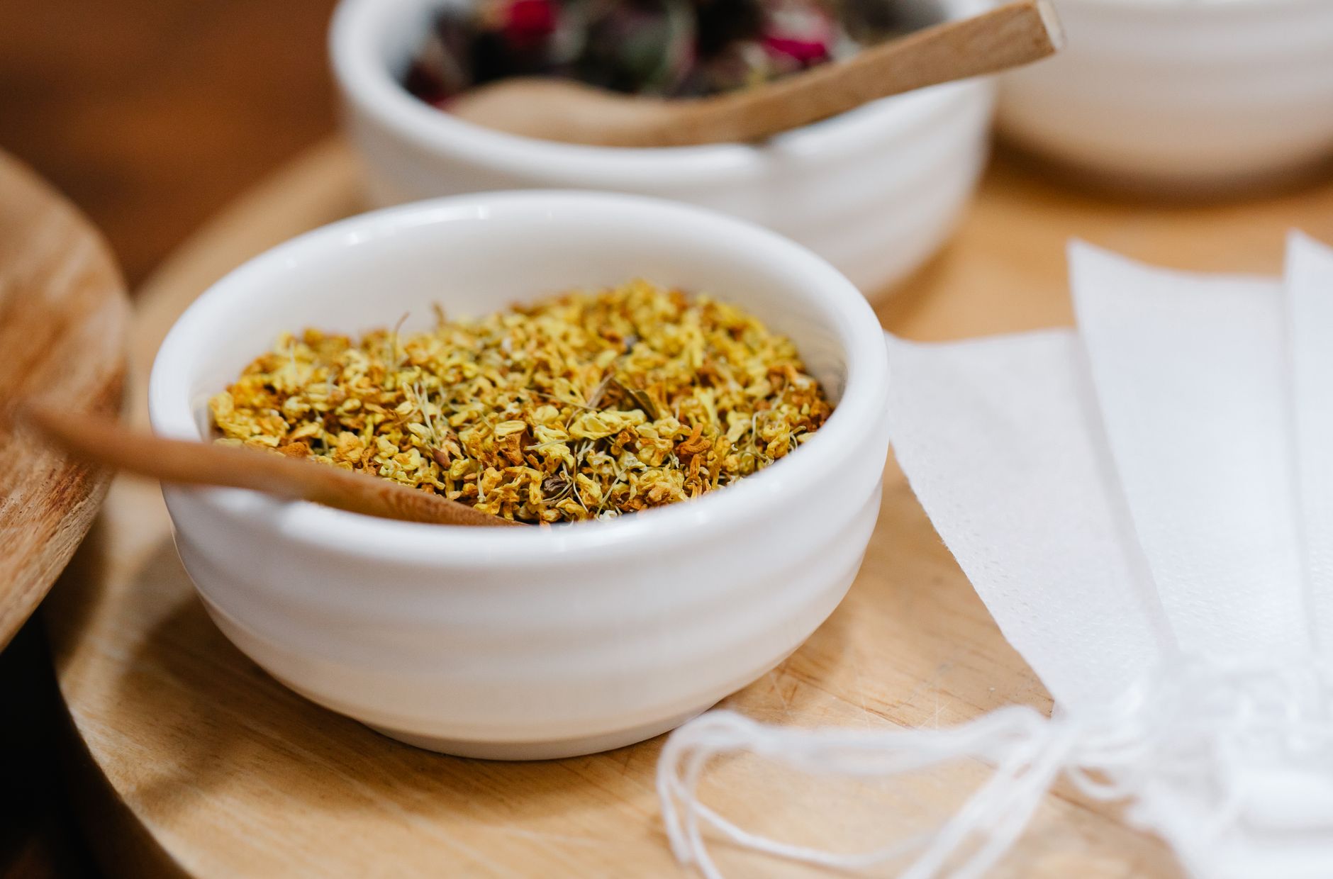 A white bowl of dried yellow flowers in front, with a wooden spoon across the rim, on a round wooden board. A second white bowl of darker dried herbs is behind it, and a folded white cloth lies to the right.