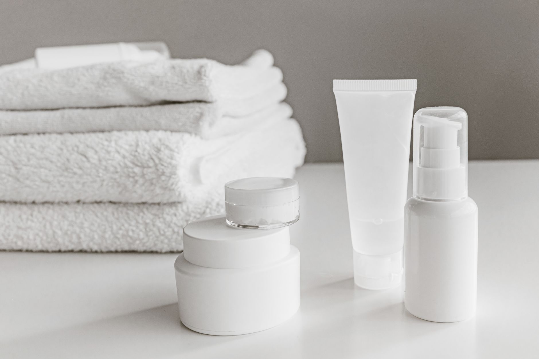 A stack of white towels beside an unlabelled white cream tube, a pump bottle and two white jars on a white surface.
