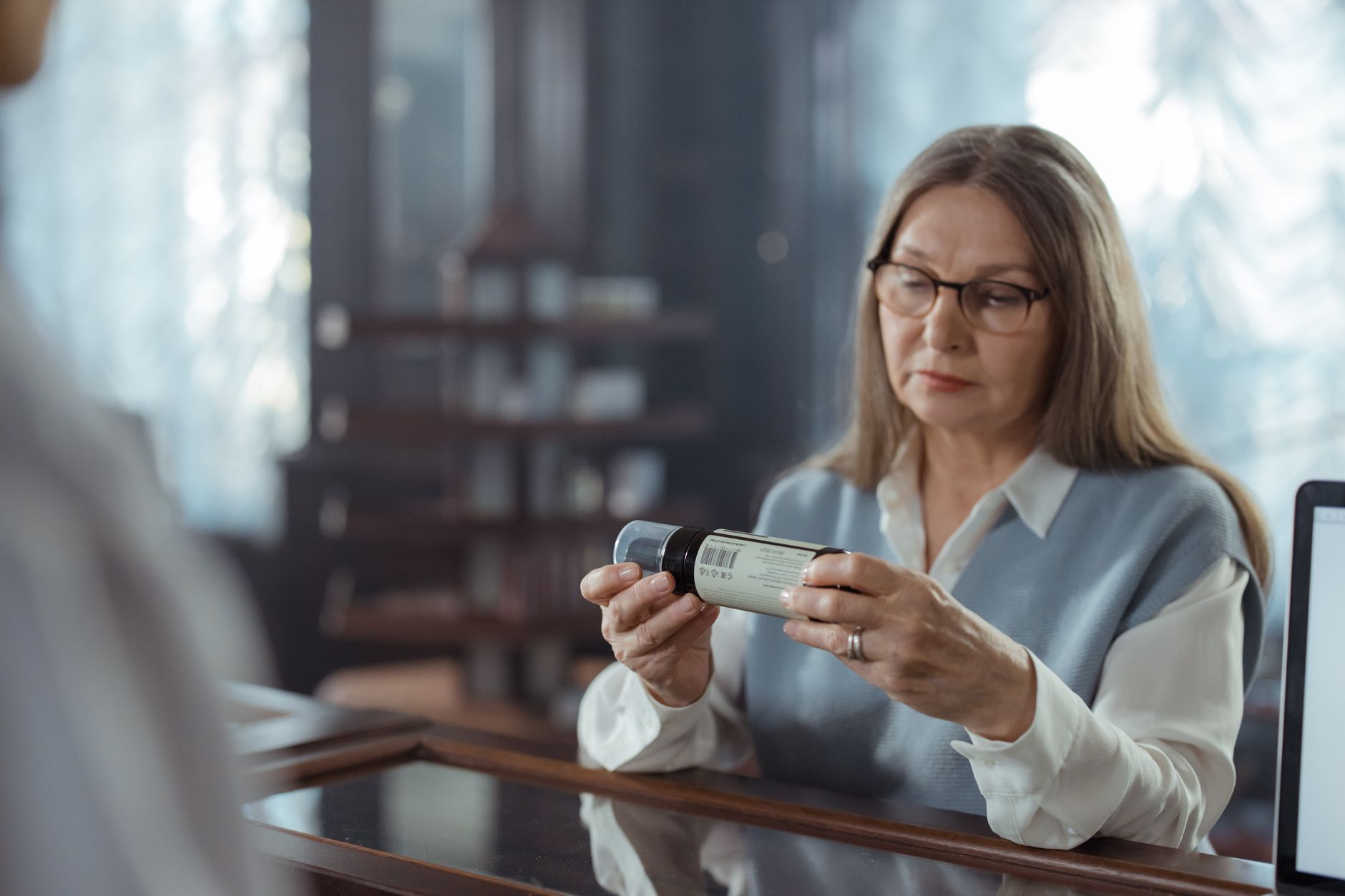 A woman in her fifties with glasses reading the label on a medicine bottle at a pharmacy counter