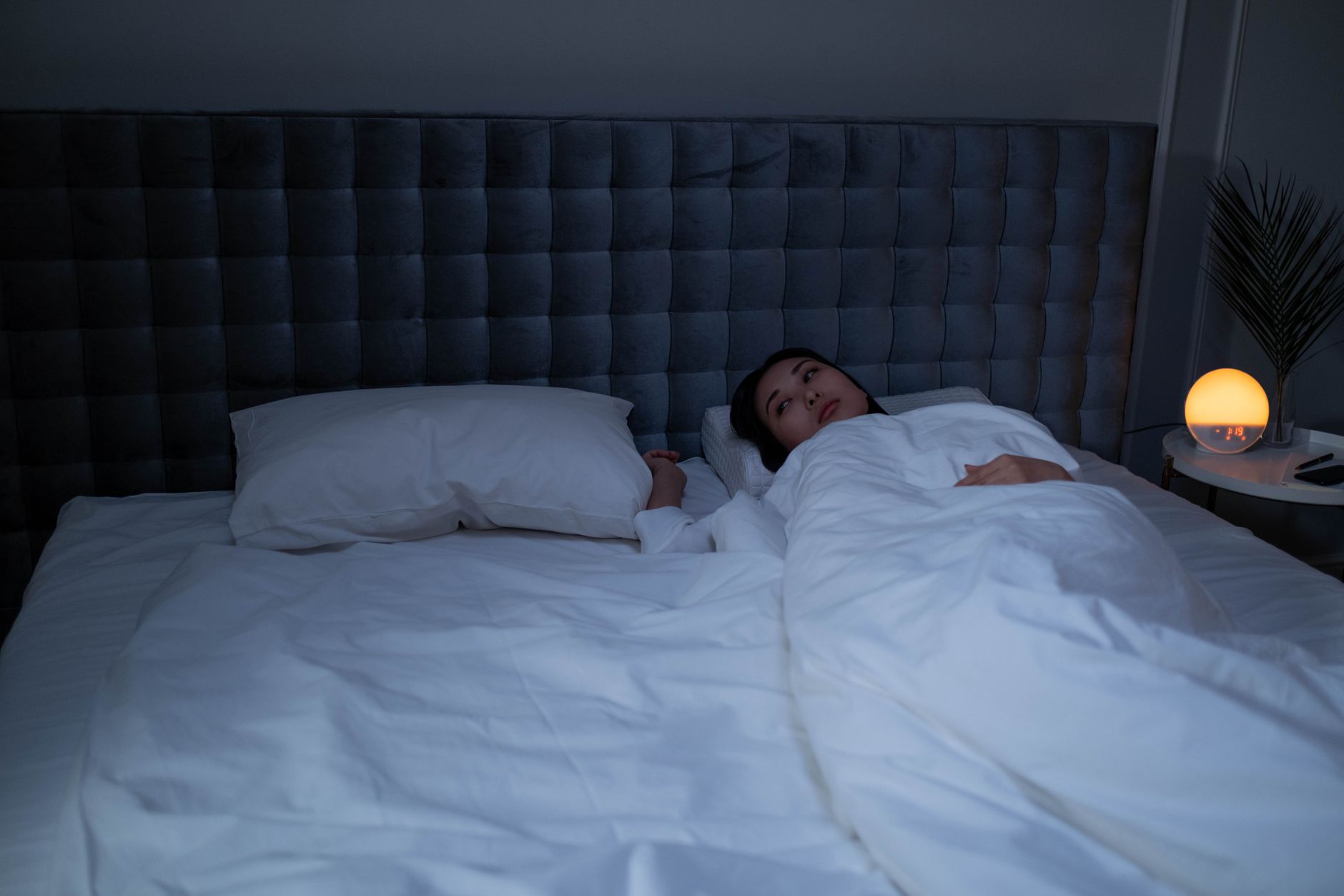 A woman lying awake under white bedding in a dim bedroom, a small round lamp glowing on the bedside table