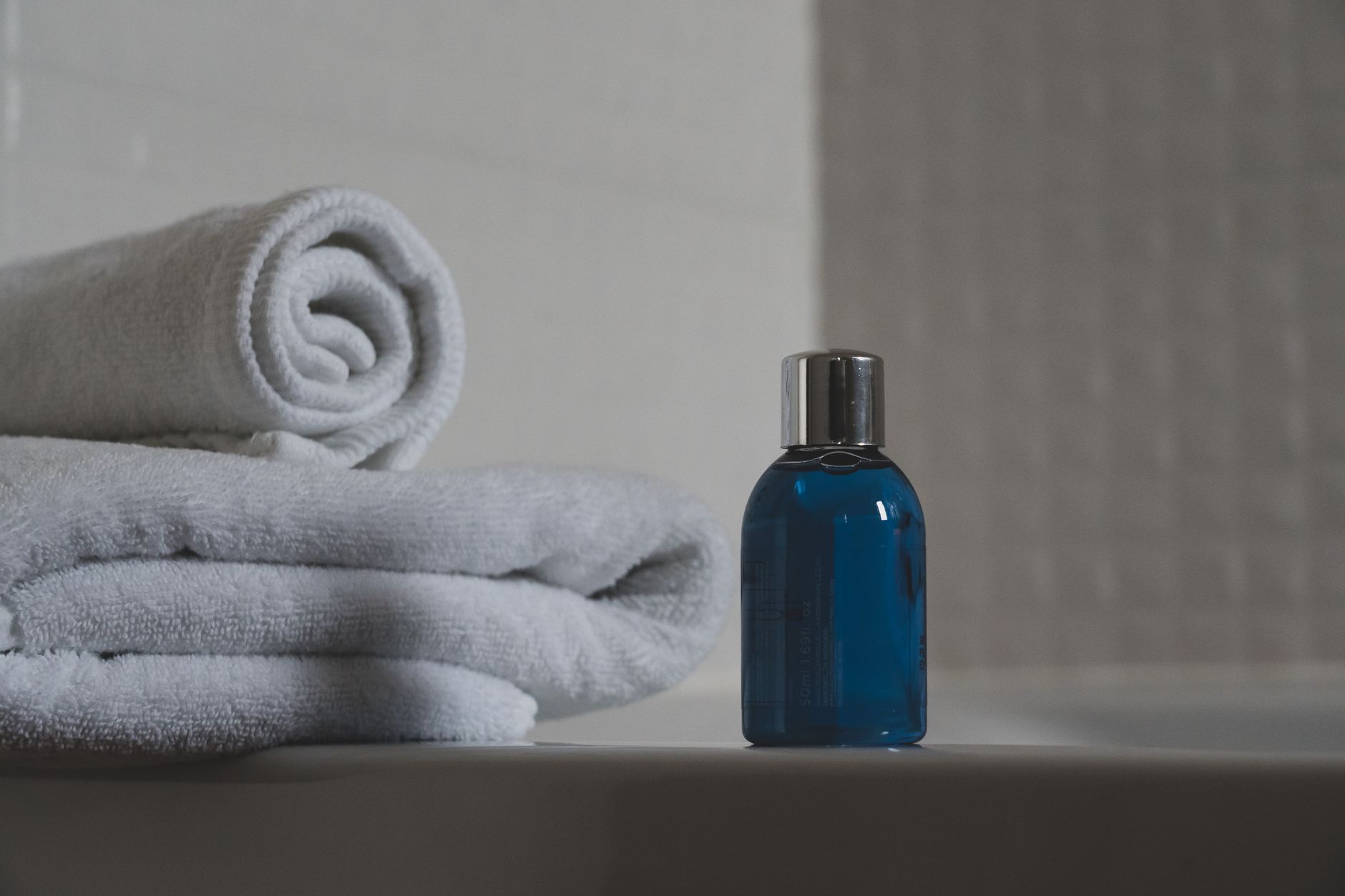 Rolled white towels beside a small blue glass bottle with a silver cap, on a pale bathroom ledge.