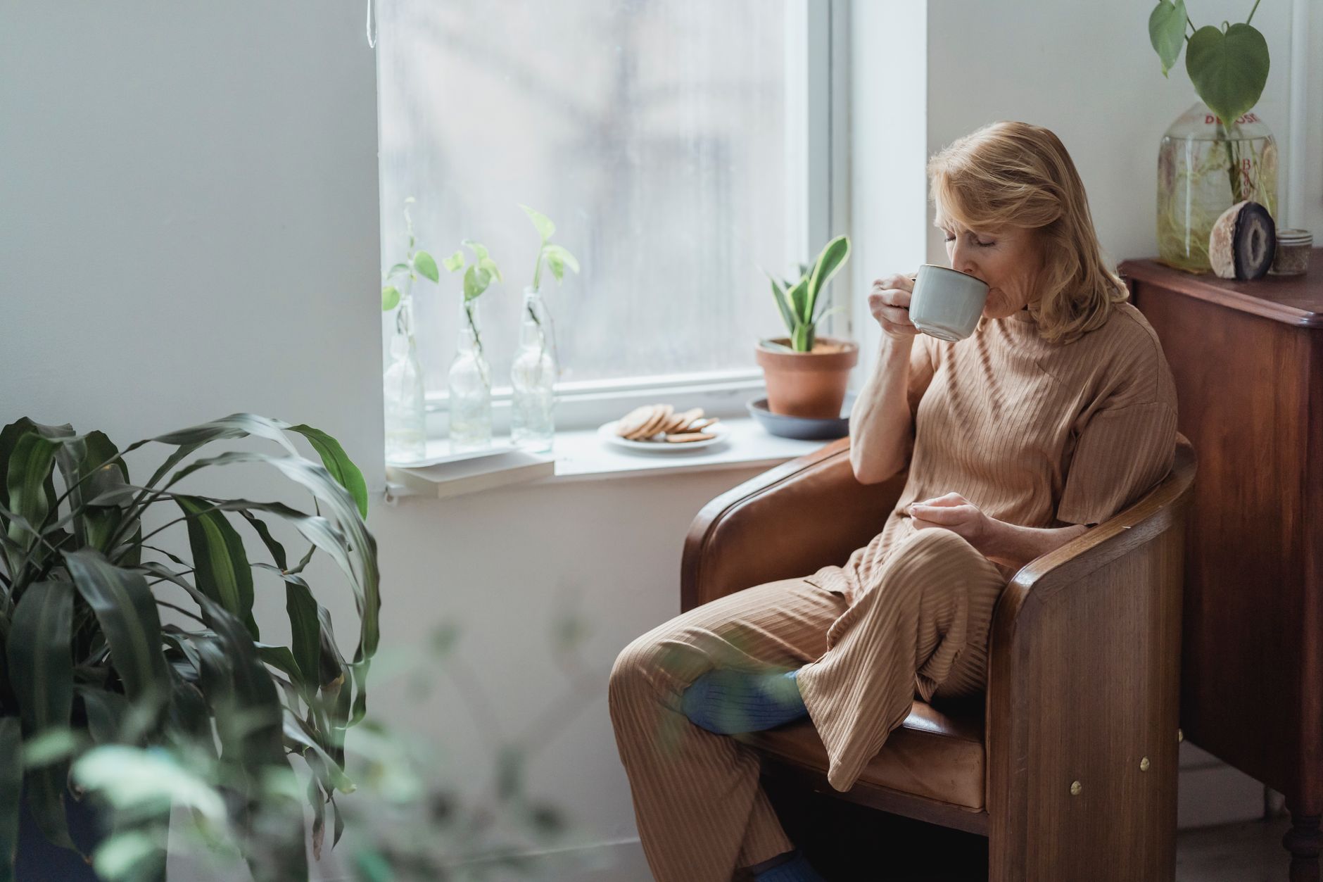 A blonde woman in a beige ribbed outfit sits in a wooden armchair beside a bright window, holding a grey mug, with houseplants and a plate of biscuits on the sill.
